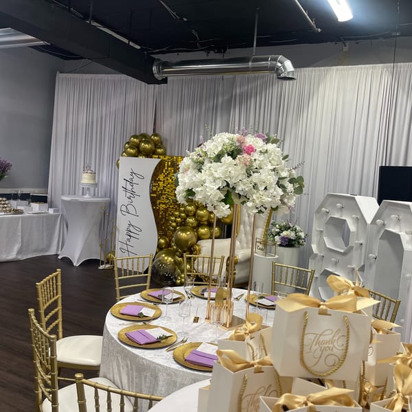 Elegant birthday party setup with gold chairs, white floral centerpieces, and "Thank You" gift bags.