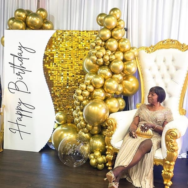 Woman in a gold dress sitting on a white and gold throne chair next to gold balloons.