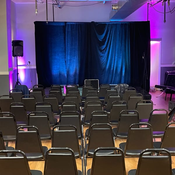 Empty black chairs face a stage with blue and purple lighting and a black curtain backdrop.