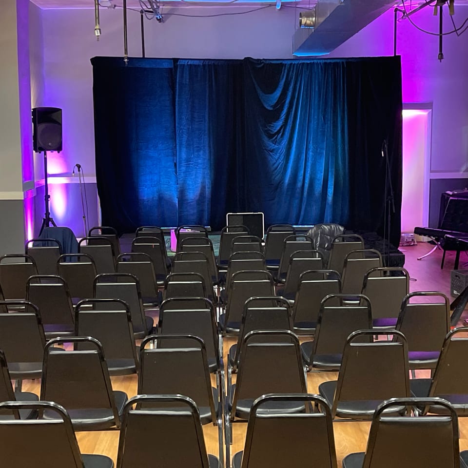 Empty black chairs face a stage with blue and purple lighting and a black curtain backdrop.