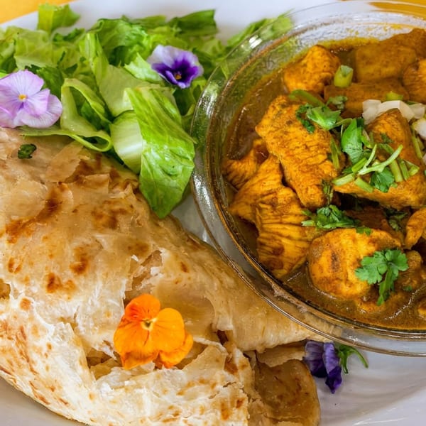 Chicken curry with paratha and a side salad garnished with edible flowers on a white plate.