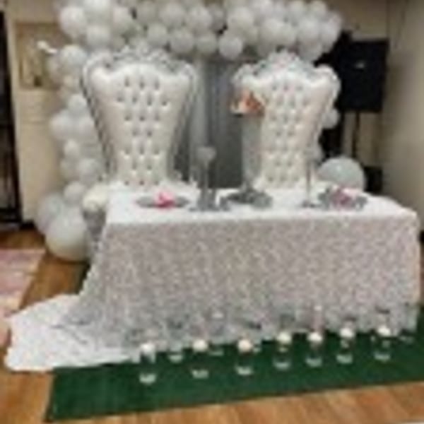 Two white tufted chairs behind a lace-covered table with white balloons and candles.