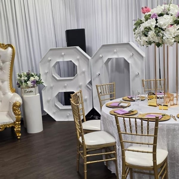Elegant event space with white draping, large illuminated "88" numbers, and a set table with gold chairs.